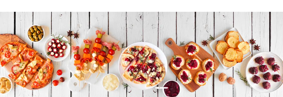 Overhead view table scene on a white wood banner background. Flatbreads, skewers, cranberry crostini and a variety of party food.