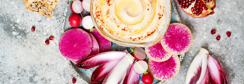 Homemade hummus seasoned with olive oil and paprika and fresh vegetables: radishes, watermelon radish, red chicory, pomegranate.