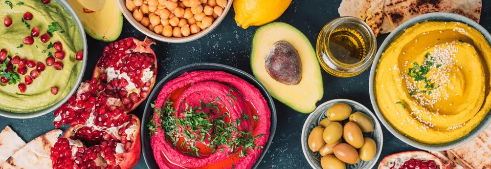 Ingredients for yellow turmeric hummus, green parsley hummus and red beetroot hummus on dark background, lemon, sesame, olive oil, pita, avocado, pomegranate.